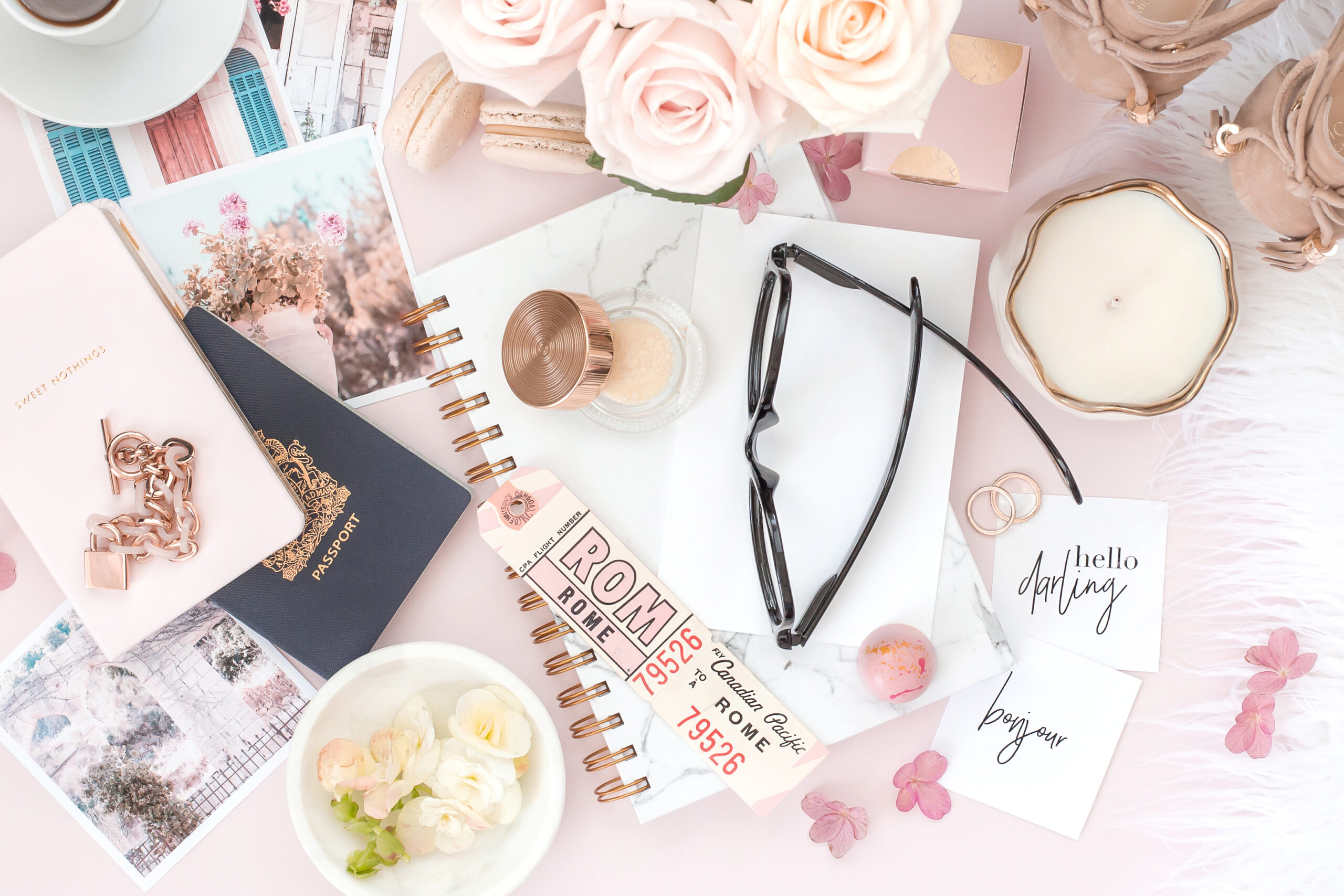 flatlay of items including a journal, a passport, glasses, and pink flowers