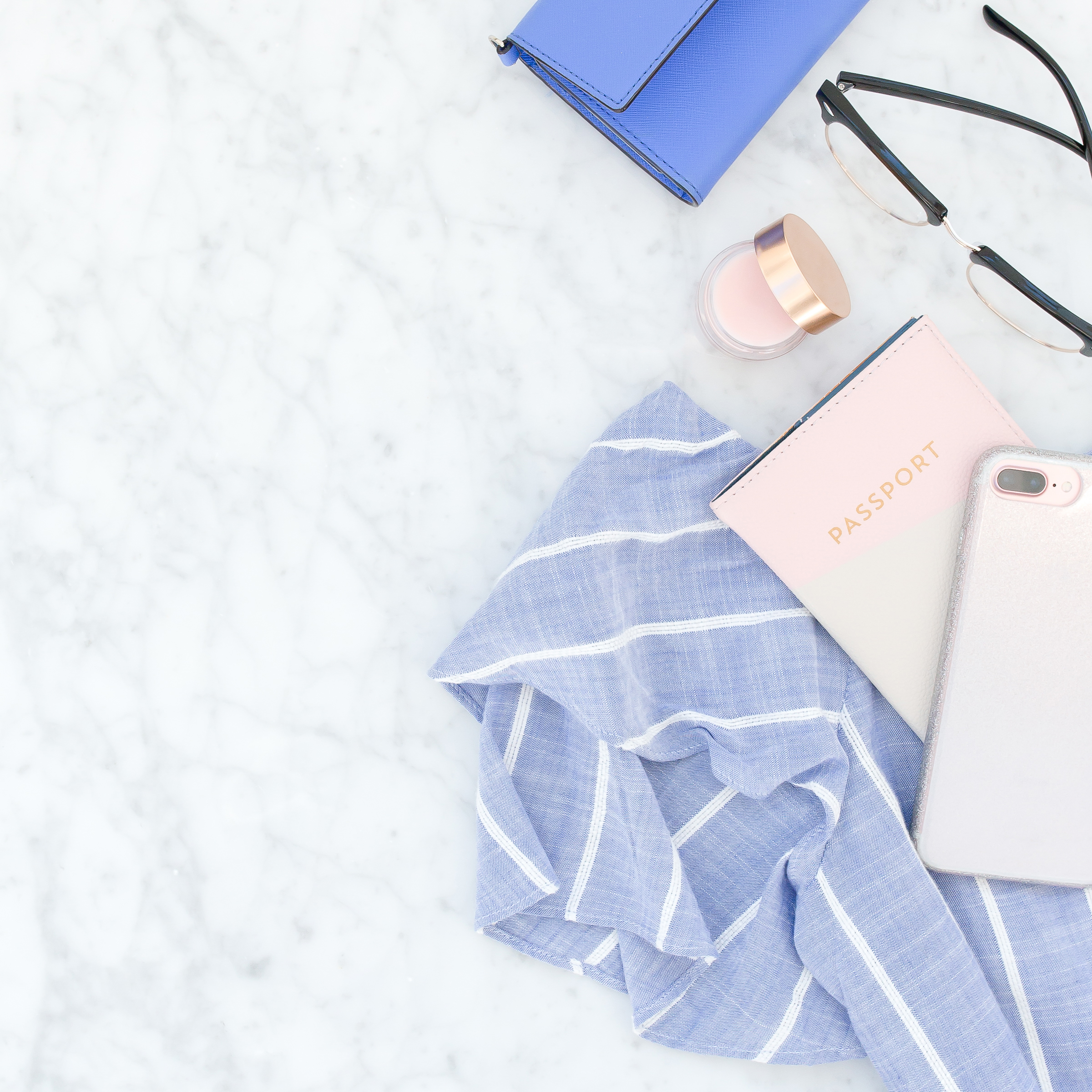 flat lay style image with a blue striped shirt, pink passport, an iphone, glasses, and a few other pink and blue accessory items